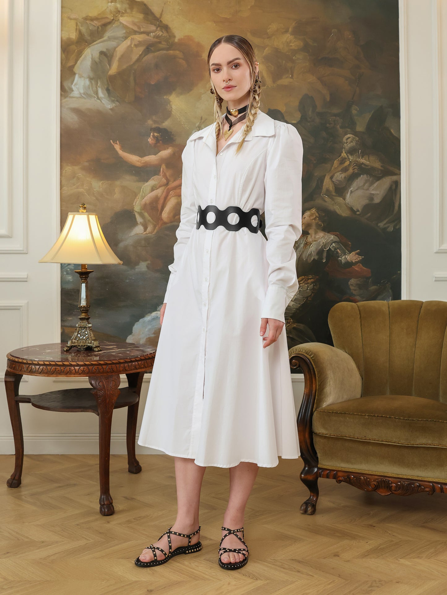 Wide black leather belt with circular cutouts worn around the waist of a button-down white dress.