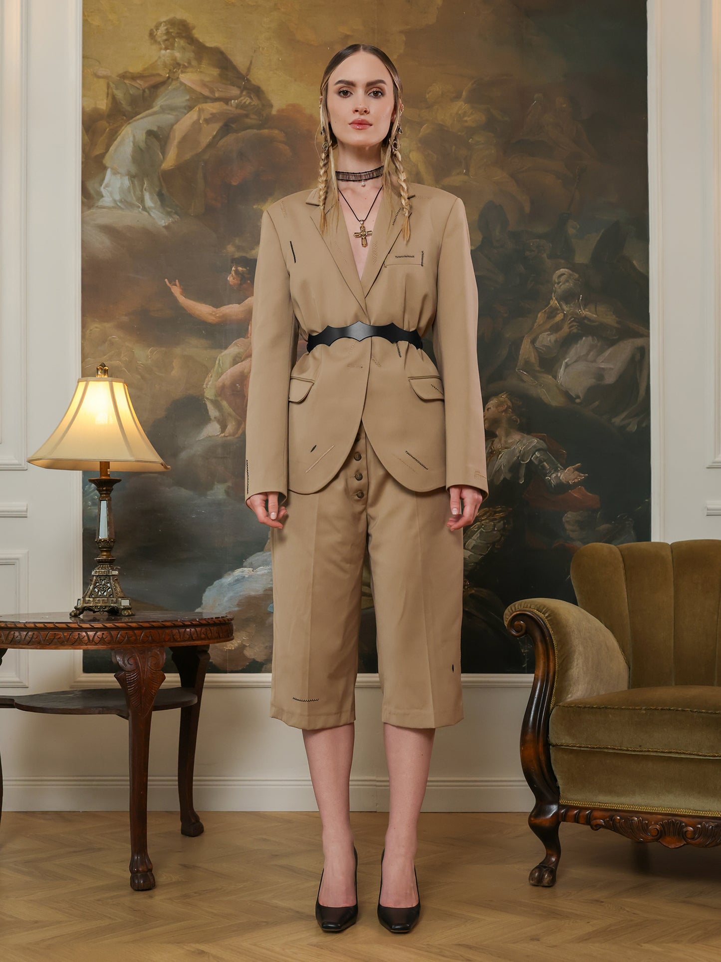 Full-body view of a woman in a beige suit and cropped trousers, styled with a black leather waist belt and heels.