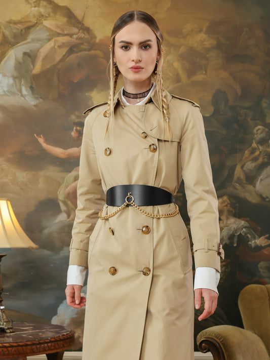 A woman in a beige trench coat styled with a wide black leather belt featuring a gold chain draped across the front.