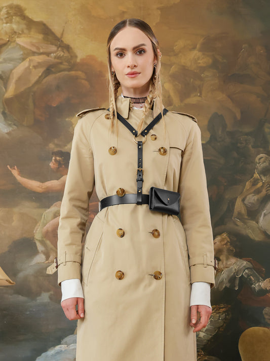 Front view of a beige trench coat accessorized with a black leather harness belt with gold hardware and detachable micro bag.