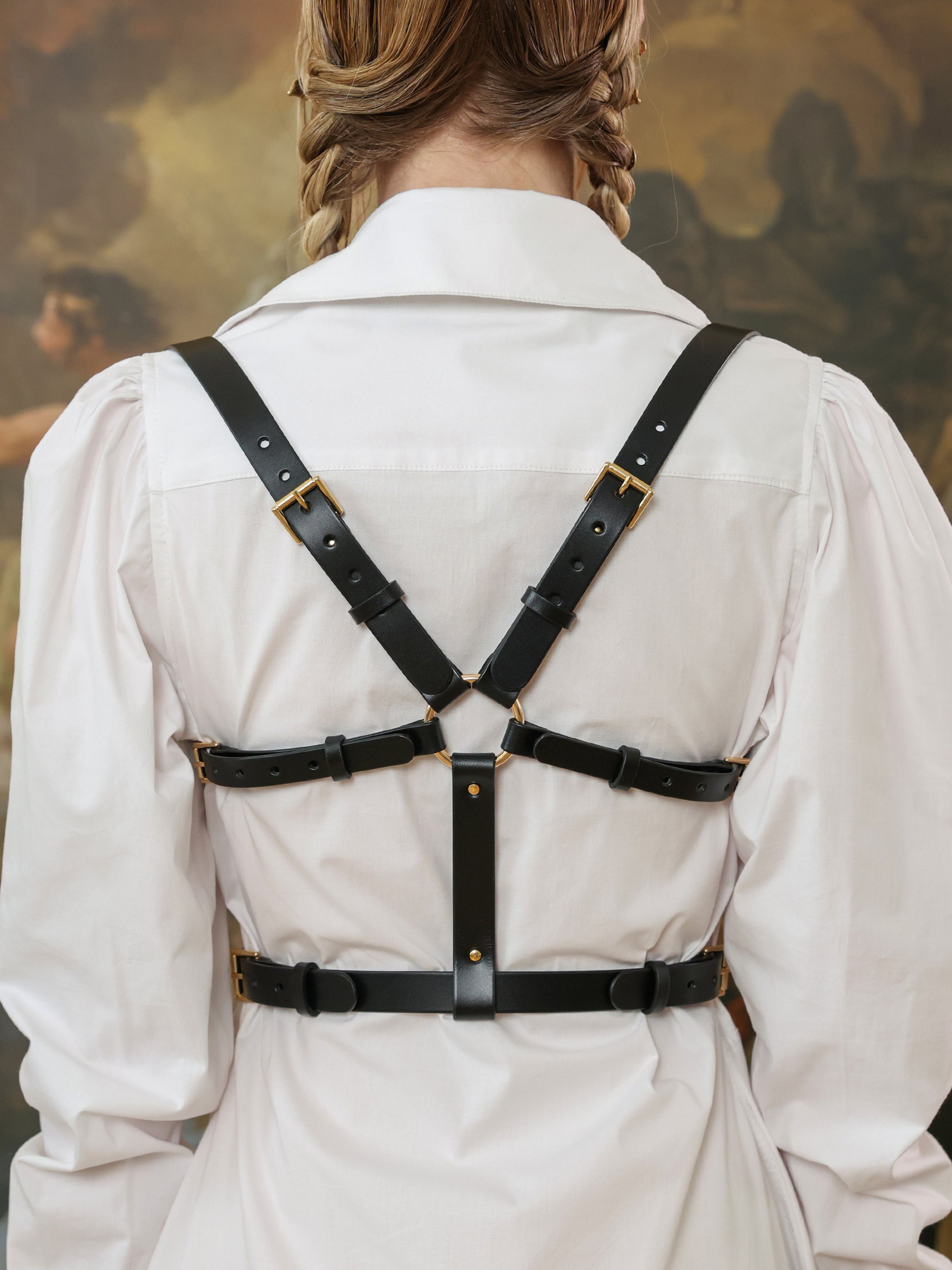 Woman wearing a white shirt dress styled with a black leather underbust harness, back view showing adjustable straps with gold buckles.