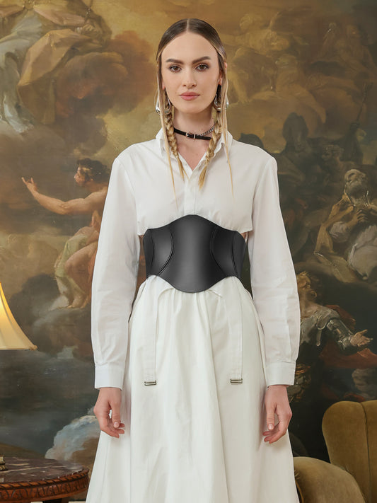 A woman wears a structured black leather corset belt over a white shirt dress, accentuating the waist with a sculpted silhouette.