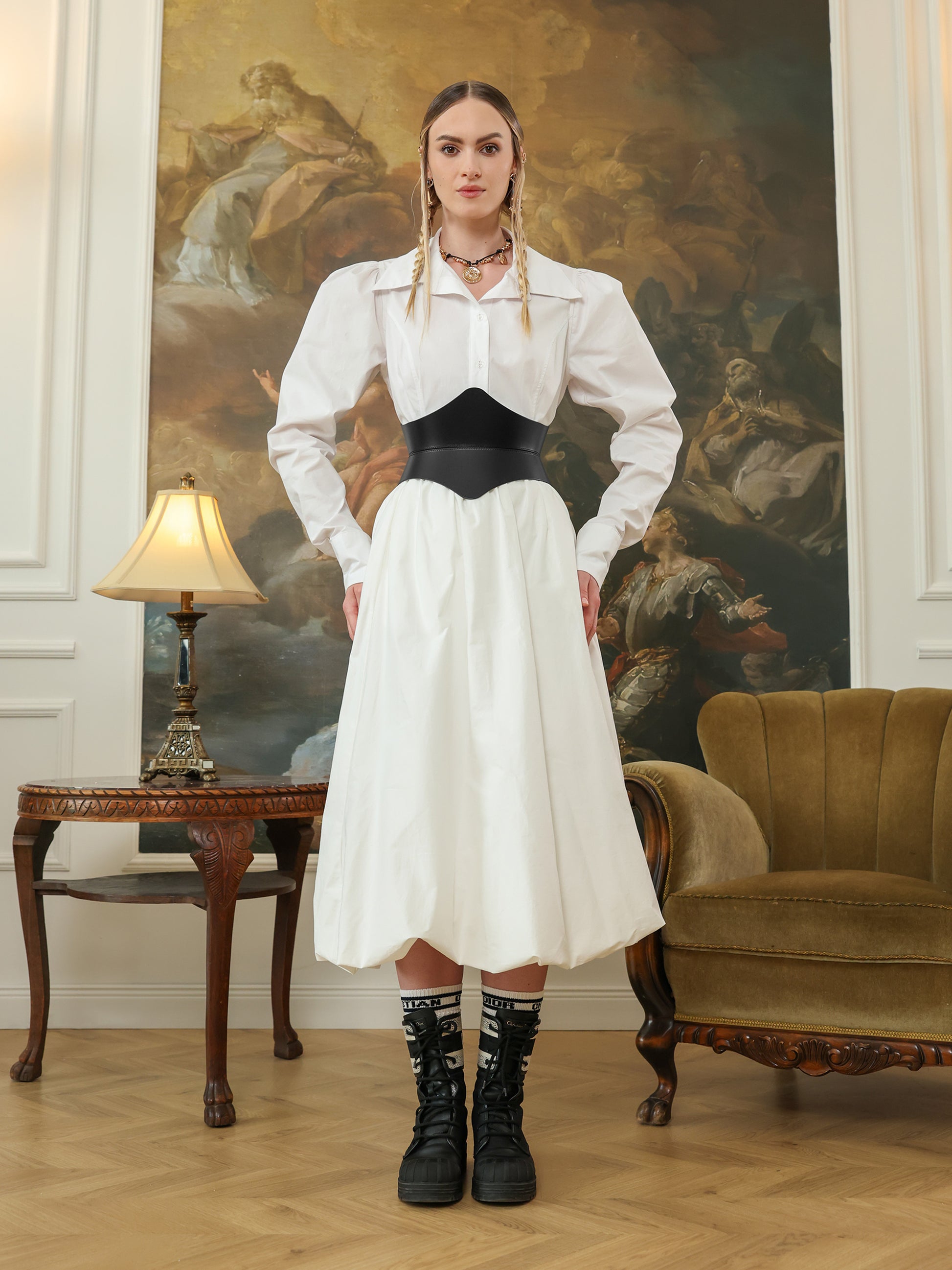 A woman in a white dress shirt and skirt wearing a black leather hourglass corset belt, paired with black lace-up boots and statement socks.