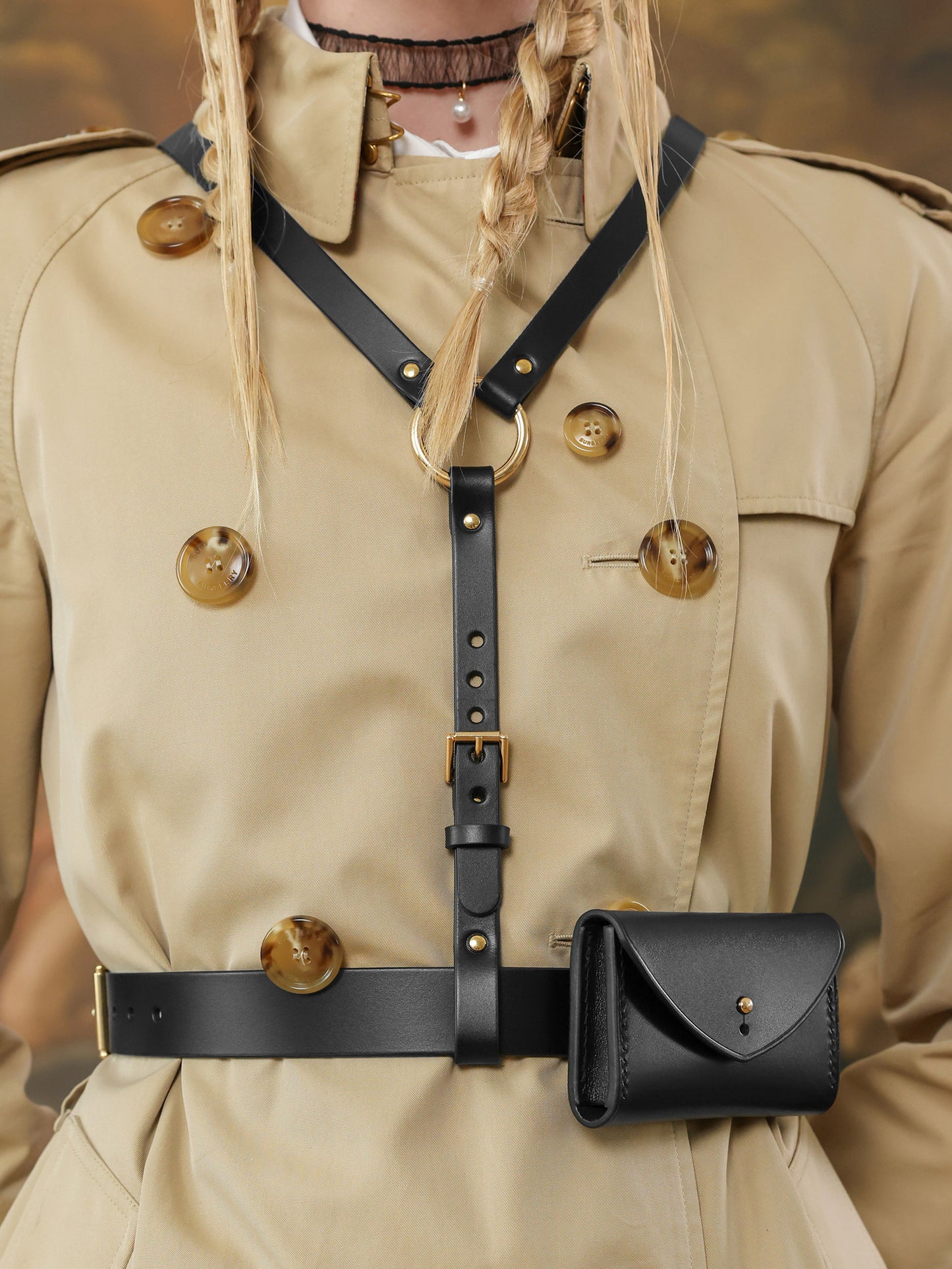 Close-up of a black leather harness belt with gold buckle and small structured micro bag on a beige trench coat.