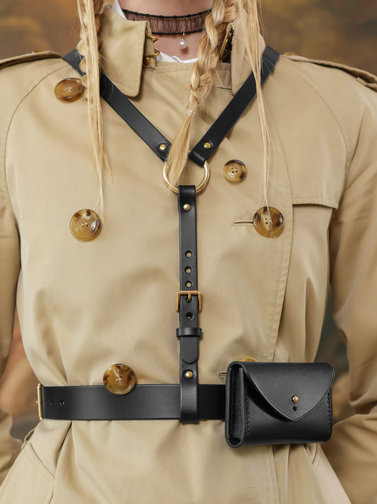 Close-up of a black leather harness belt with gold buckle and small structured micro bag on a beige trench coat.