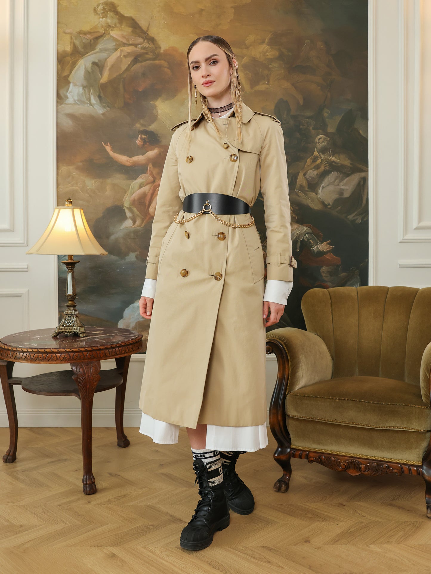 Full outfit view of a woman in a beige trench coat and boots, styled with a wide black leather belt with gold chain detail.