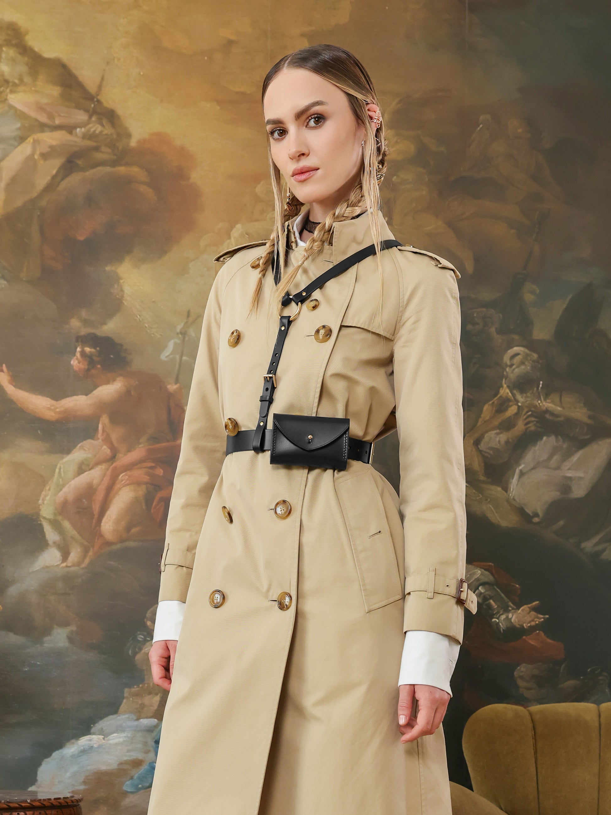 Side view of a beige trench coat styled with a black leather harness featuring a micro bag at the waist.
